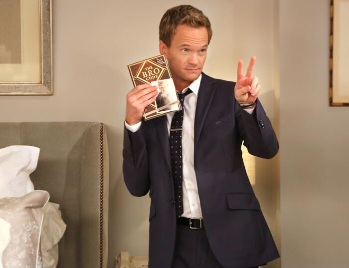 Man in a dark suit holding a book titled The Bro Code, making a peace sign, related to celebrities people used to like then hate.