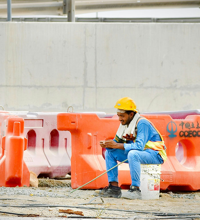 Construction worker wearing a helmet resting on a bucket at a dusty labor camp site in Dubai with orange barriers.