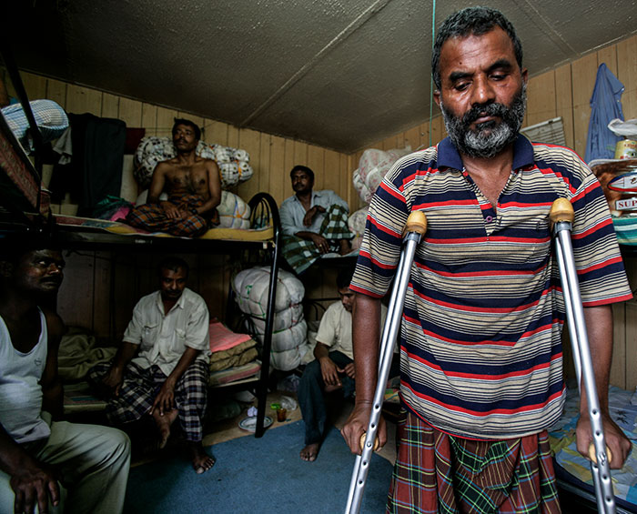 Man with crutches and striped shirt in crowded room with other men, depicting harsh reality of Dubai labor camps conditions.