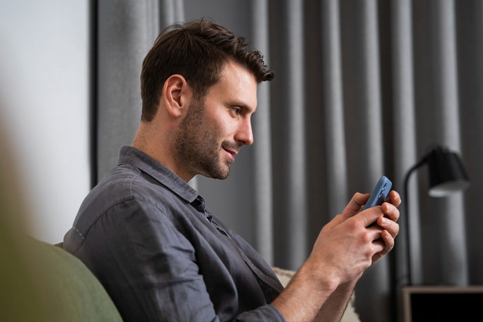 Man smiling and texting on phone while seated indoors, capturing a moment related to a date ambush scenario.