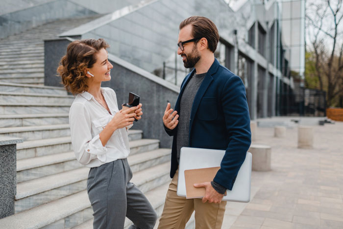 Woman hypes up stunner friend to single coworker, smiles while holding a ring box during an outdoor conversation near stairs.