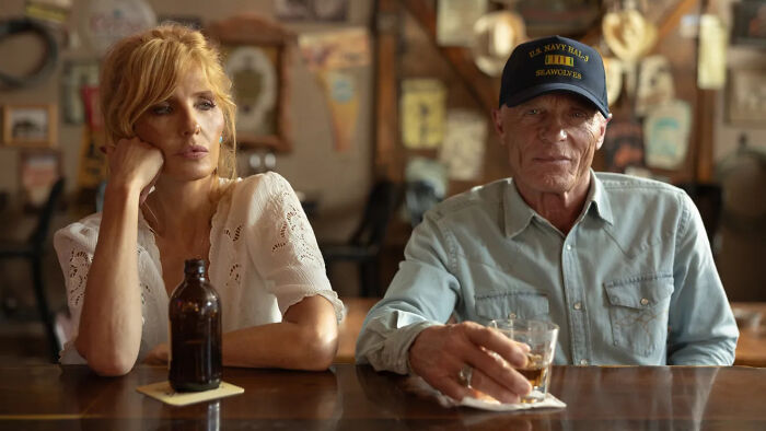 Two people sitting at a bar, a woman looking pensive and a man wearing a hat holding a drink, relating to Dutton Ranch.