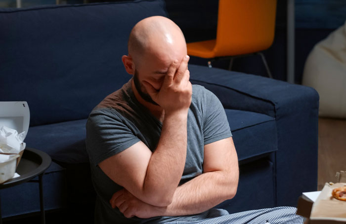 Man sitting on floor with head in hand, upset after eating bigger share of sandwich at casual party setting.