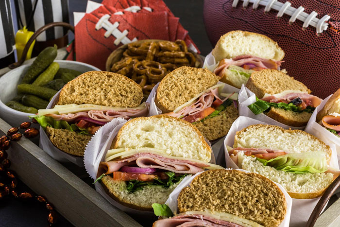 Tray of sandwiches with lettuce and ham served with pickles and pretzels at a sandwich party setting with footballs.
