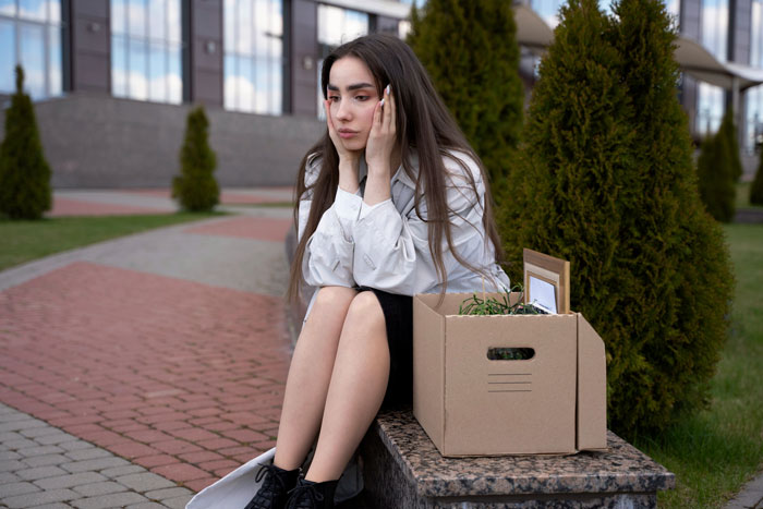 Young woman sitting outside with a cardboard box, looking upset after emailing roommates about boss stealing portfolio.