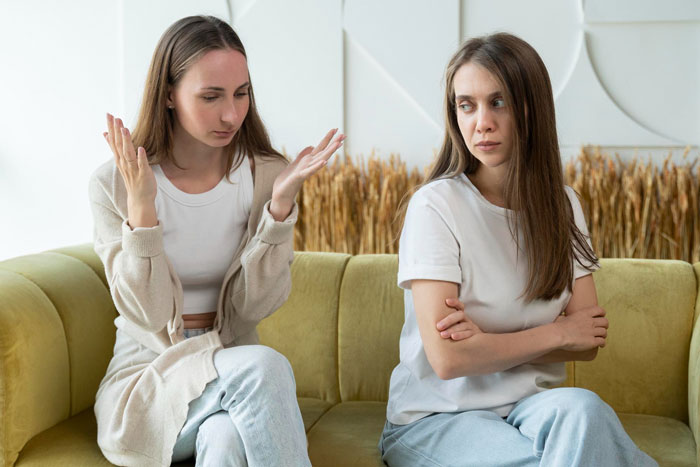 Two women on a couch having a tense conversation about emailing roommates and a boss who stole a portfolio.