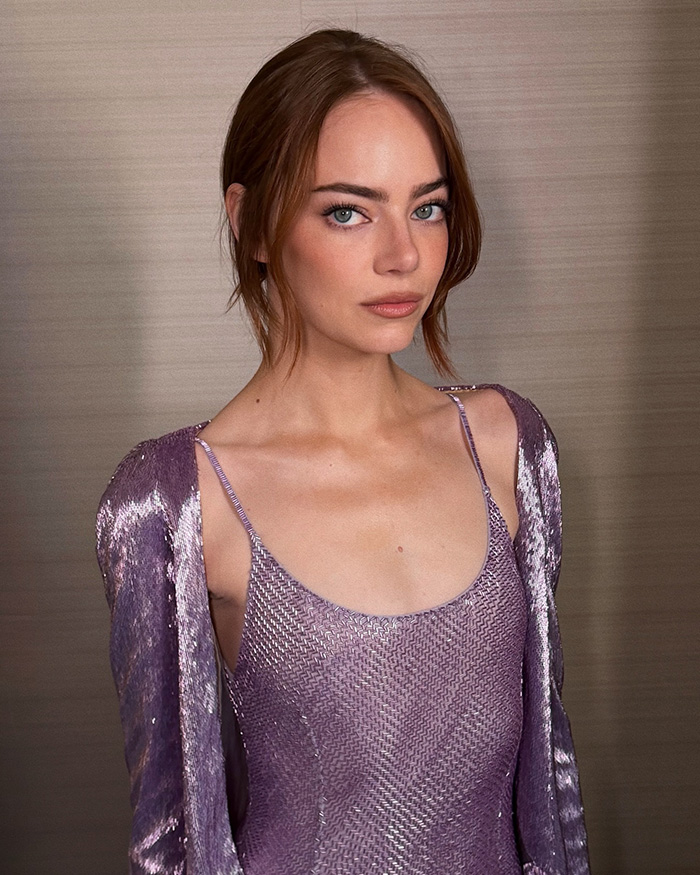 Emma Stone in a shimmering purple dress and matching jacket, photographed indoors with a neutral background.
