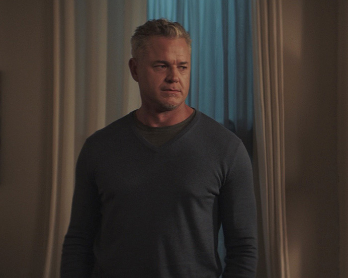 Eric Dane wearing a dark sweater standing indoors near curtains, reflecting on the cause of passing revealed recently.