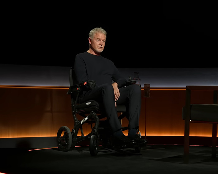 Man in black sitting in a wheelchair on a dark stage with dim orange lighting, relating to Eric Dane&rsquo;s cause of passing.