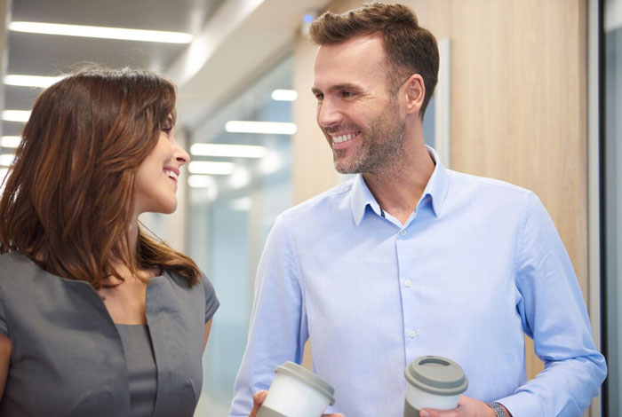 Two colleagues smiling and chatting in an office hallway, woman showing confidence as a cancer survivor at work.