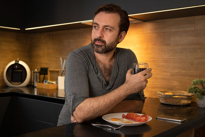 Man in kitchen holding glass, looking pensive about family issues and regrets related to wedding and father cutting off family.