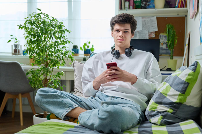 Young man sitting on bed with phone and headphones, reflecting on family issues related to dad cutting off family ties.