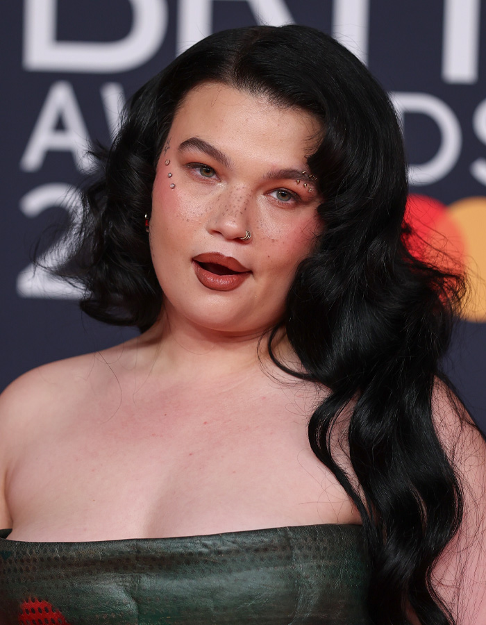 Lola Young with long black hair wearing a strapless dress resembling an oil container on a red carpet event. Lola Young with long black hair wearing a strapless dress resembling an oil container on a red carpet event.