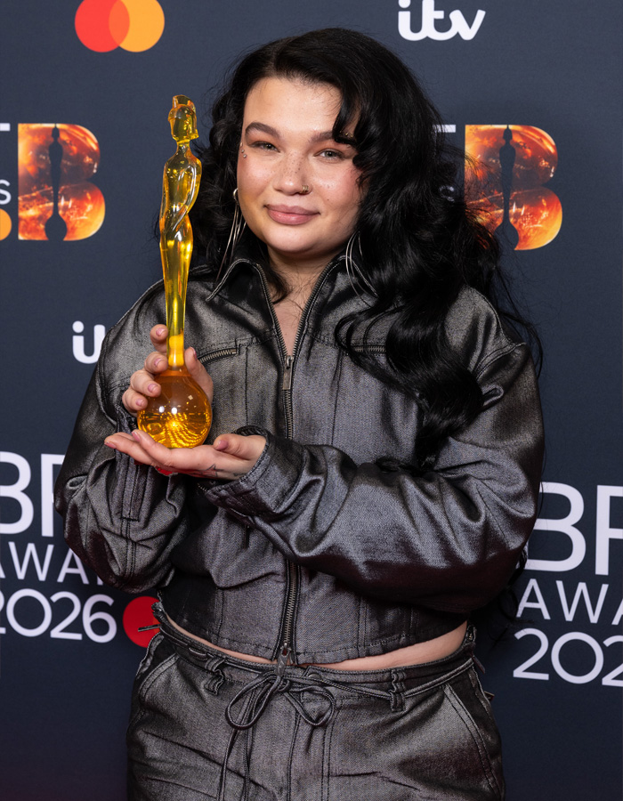 Lola Young holding a golden award statue wearing a shiny metallic outfit at the Brit Awards 2026 red carpet. Lola Young holding a golden award statue wearing a shiny metallic outfit at the Brit Awards 2026 red carpet.