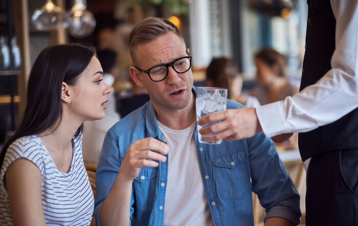 Couple at restaurant reacting with disbelief to waiter serving a glass of water, illustrating ridiculous customer complaints.