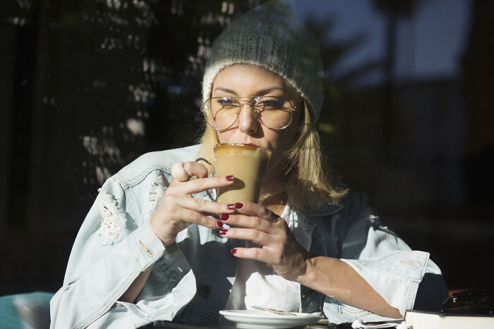 Woman wearing glasses and a knit hat sipping coffee in a cafe, illustrating customers’ ridiculous complaints humor.
