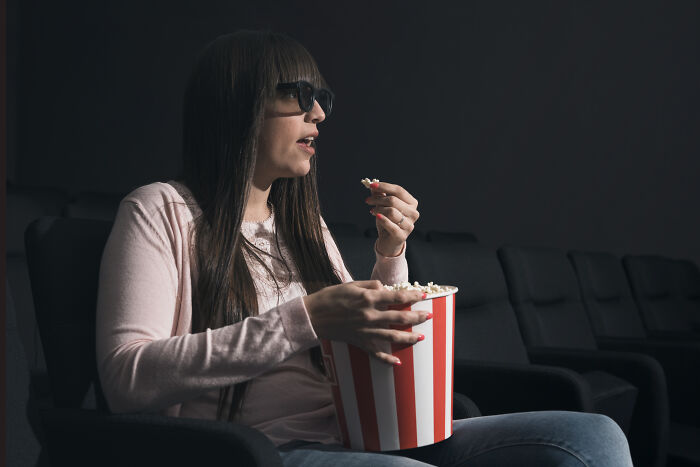 Woman wearing 3D glasses eating popcorn alone in a dark theater, illustrating customers’ ridiculous complaints humor.