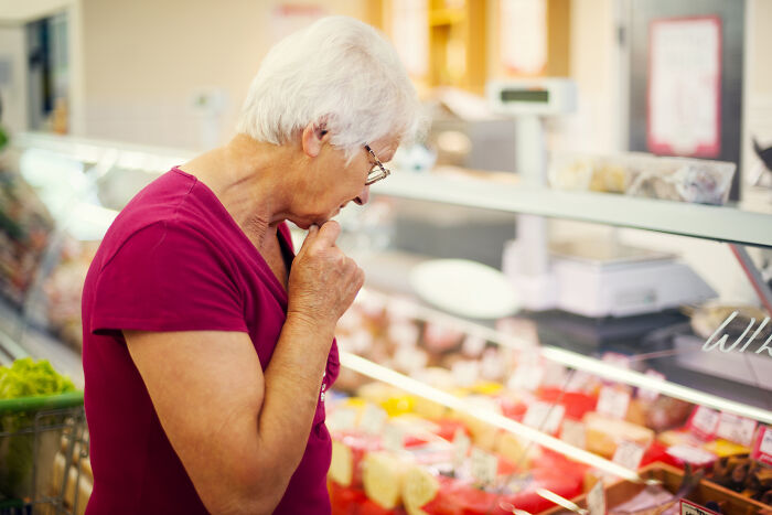 Elderly woman in a red shirt looking thoughtfully at products in a store, reflecting on customers’ ridiculous complaints.