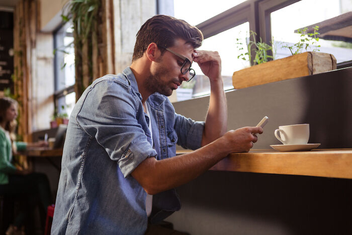 Man in denim shirt looking frustrated while reading customer complaints on phone in a cafe setting with coffee cup nearby