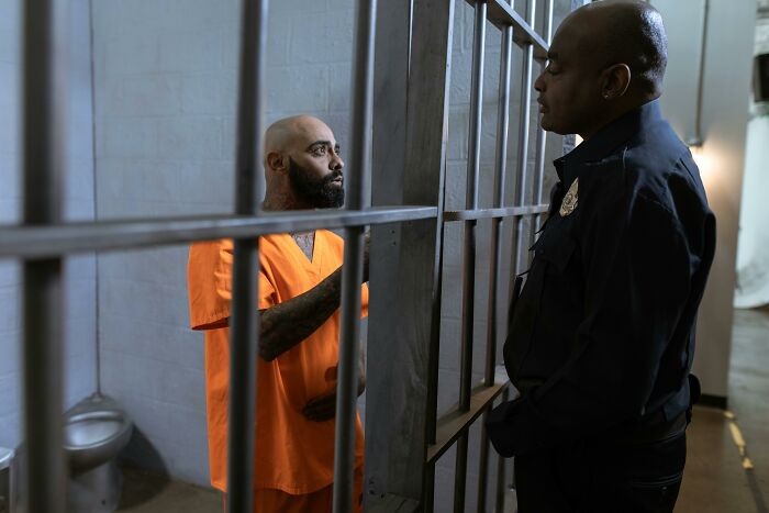 Inmate in orange prison uniform talking to guard behind bars, illustrating stories of personal hell and ruined lives.
