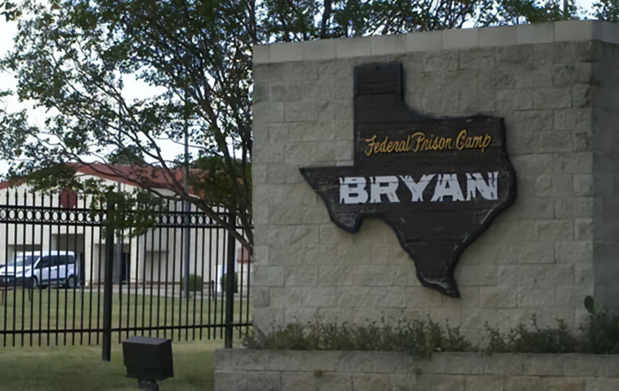 Sign reading Federal Prison Camp Bryan mounted on stone wall outside the gated prison facility in Texas on a clear day.