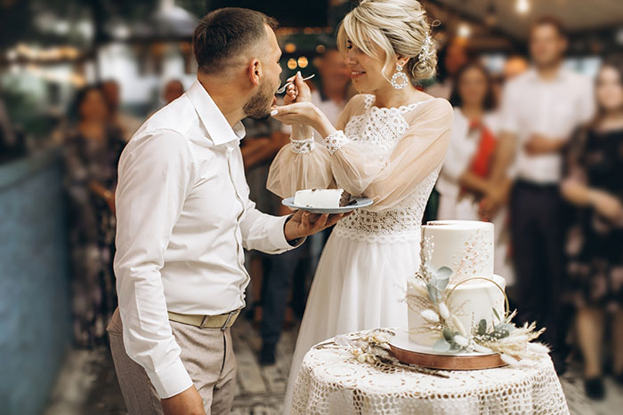 Noiva de vestido branco alimentando bolo de noivo na recepção de casamento com convidados comemorando ao fundo