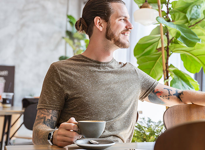 Man with tattoos holding coffee cup and smiling by window in cafe, illustrating first dates character-building trauma stories.