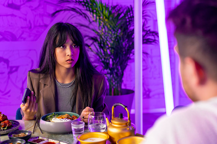 Young woman looking uneasy on a first date at a restaurant with colorful lighting, experiencing character-building trauma.