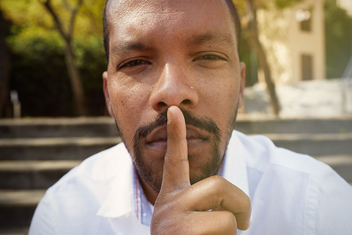 Man in white shirt with finger to lips signaling silence, reflecting on first dates so horrible they build character trauma.