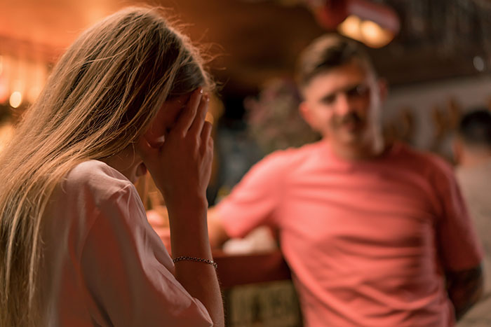 A woman covering her face in distress on a first date with a man at a dimly lit bar, showing first date trauma.