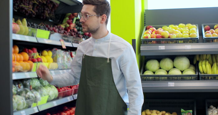 Man wearing apron arranging fresh fruits and vegetables in a grocery store focused on change and loneliness themes