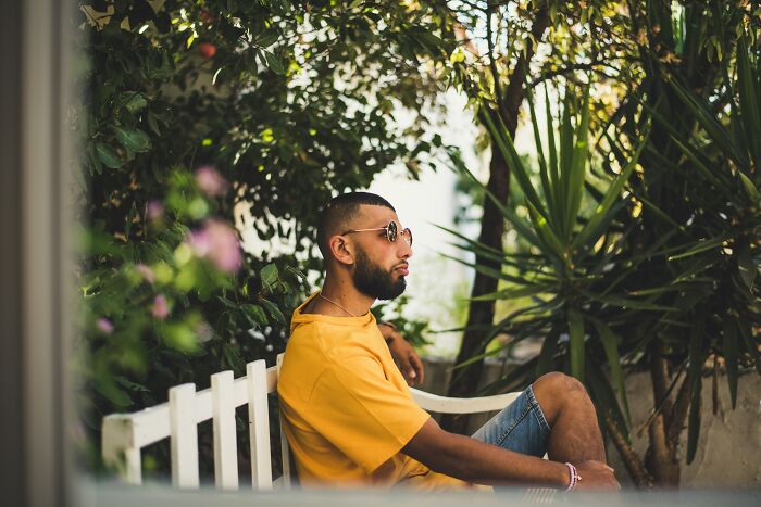 Young man wearing sunglasses and a yellow shirt sitting thoughtfully on a bench surrounded by greenery reflecting on change.