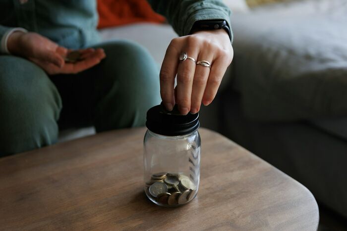 Person placing coins into a glass jar on a wooden table, symbolizing life hacks and personal savings regrets.