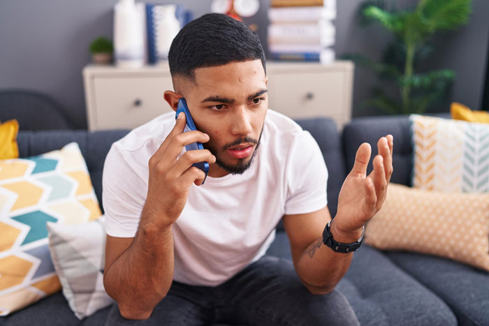 Man in a white t-shirt speaking on phone looking frustrated while sitting on a couch with patterned pillows nearby
