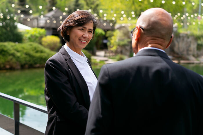 Two funeral directors in black suits having a friendly conversation outdoors near a pond with greenery in the background