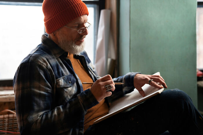 Older man wearing a red beanie and glasses, writing in a notebook while reflecting on wild things funeral directors saw.