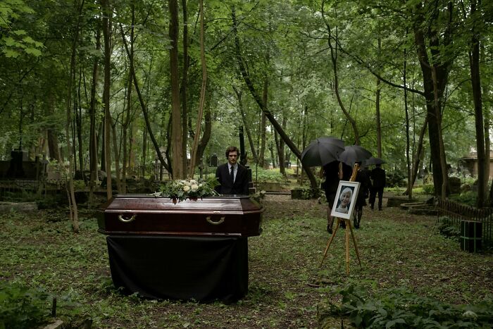 Coffin in wooded cemetery with funeral directors and attendees holding umbrellas during an outdoor funeral service.