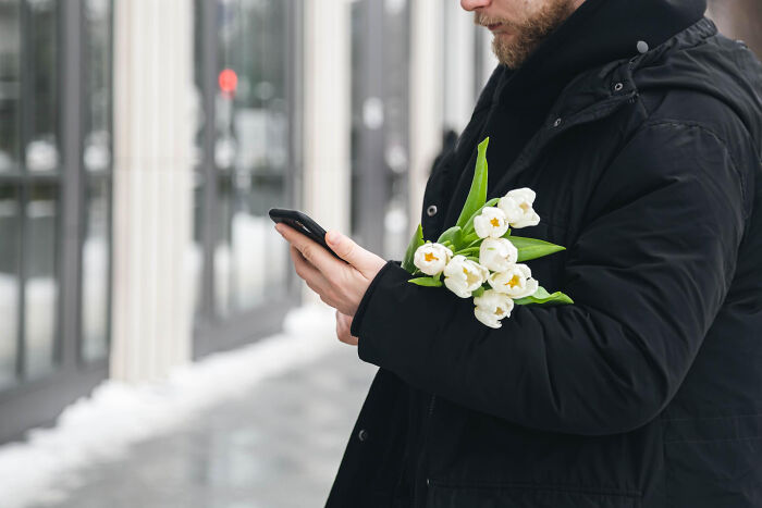 Man in black coat holding white tulips and using smartphone outside, illustrating wild things funeral directors saw moments.