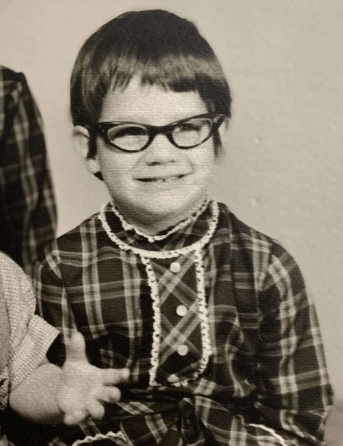 Vintage childhood photo showing a young child with a hair disaster and glasses wearing a plaid dress.