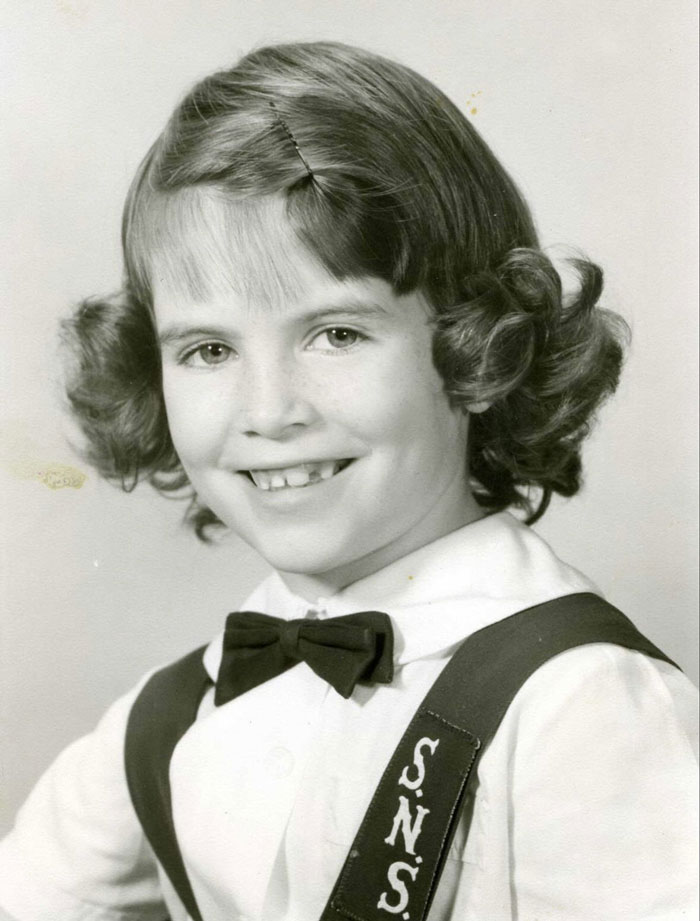 Black and white photo of a smiling child with a vintage curly hairstyle, showcasing a childhood hair disaster style.