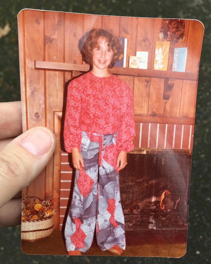 Childhood photo of a girl with a hair disaster, wearing patterned clothes, standing by a wood-paneled fireplace.