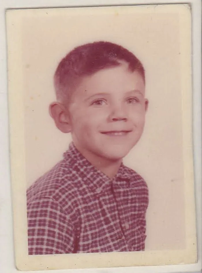Vintage childhood photo of boy with a hair disaster, wearing a checkered shirt and smiling at the camera.