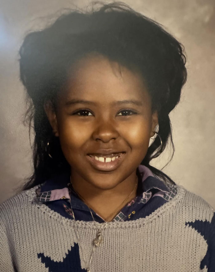 Smiling young girl from childhood with a noticeable hair disaster wearing hoop earrings and a patterned sweater.