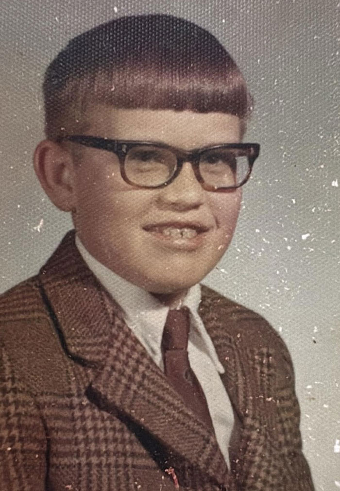 Childhood photo of a boy with a vintage hair disaster, wearing glasses and a plaid suit jacket and tie.