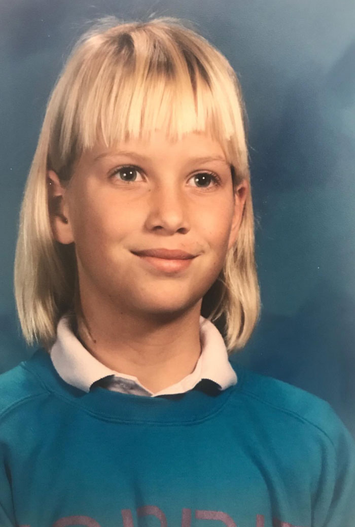 Childhood photo of a girl with a hair disaster, short uneven bangs and shoulder-length blonde hair smiling.