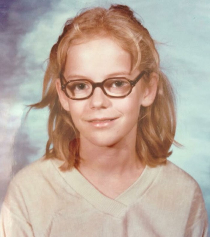 Childhood photo of a girl with a hair disaster wearing glasses and a beige sweater against a studio backdrop.