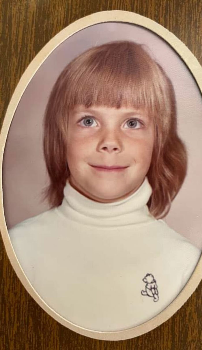 Childhood photo of a young child with a hair disaster hairstyle wearing a white turtleneck with a teddy bear logo.