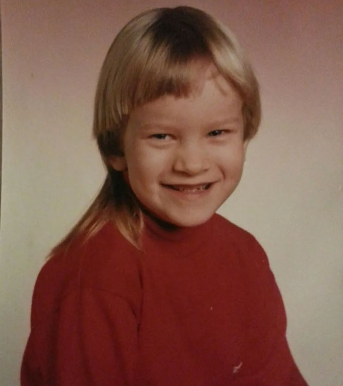 Child smiling in a vintage photo showing a childhood hair disaster with a mismatched mullet hairstyle and uneven bangs.
