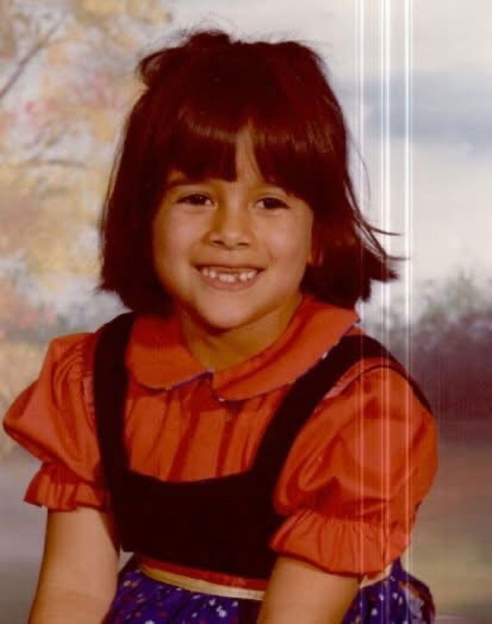 Childhood photo of a girl with a hair disaster smile, wearing an orange dress outdoors with a blurred fall background.