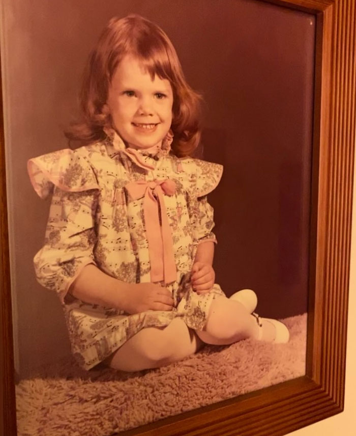 Childhood photo of a young girl smiling with a vintage hair disaster and wearing a patterned dress with a bow.